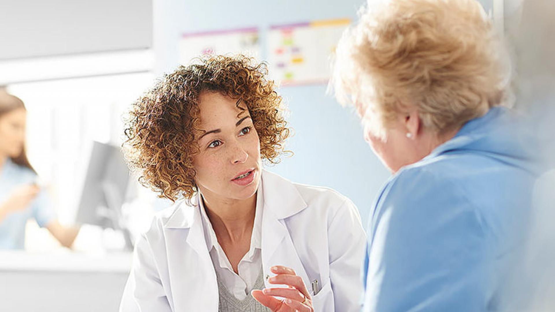 A female doctor in a white coat attentively discussing treatment options with an elderly female patient, with another medical professional in the background.