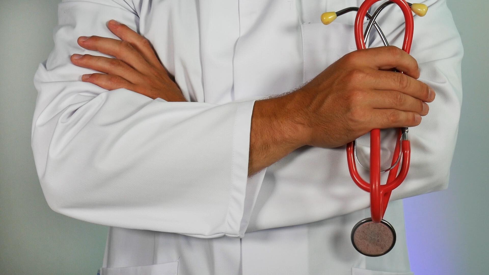 a torso shot of someone in a white doctor's coat with their arms crosses, a stethoscope in one hand