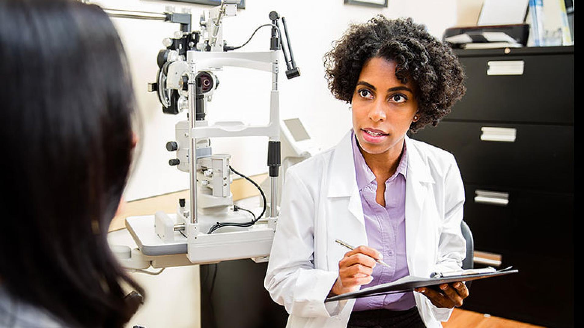 An optometrist in a white coat holds a clipboard and talks to a patient in an examination room with an eye examination machine in the background.