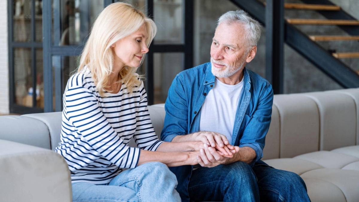 Two adults sitting close on a couch, holding hands and smiling at each other in a warmly lit living room.