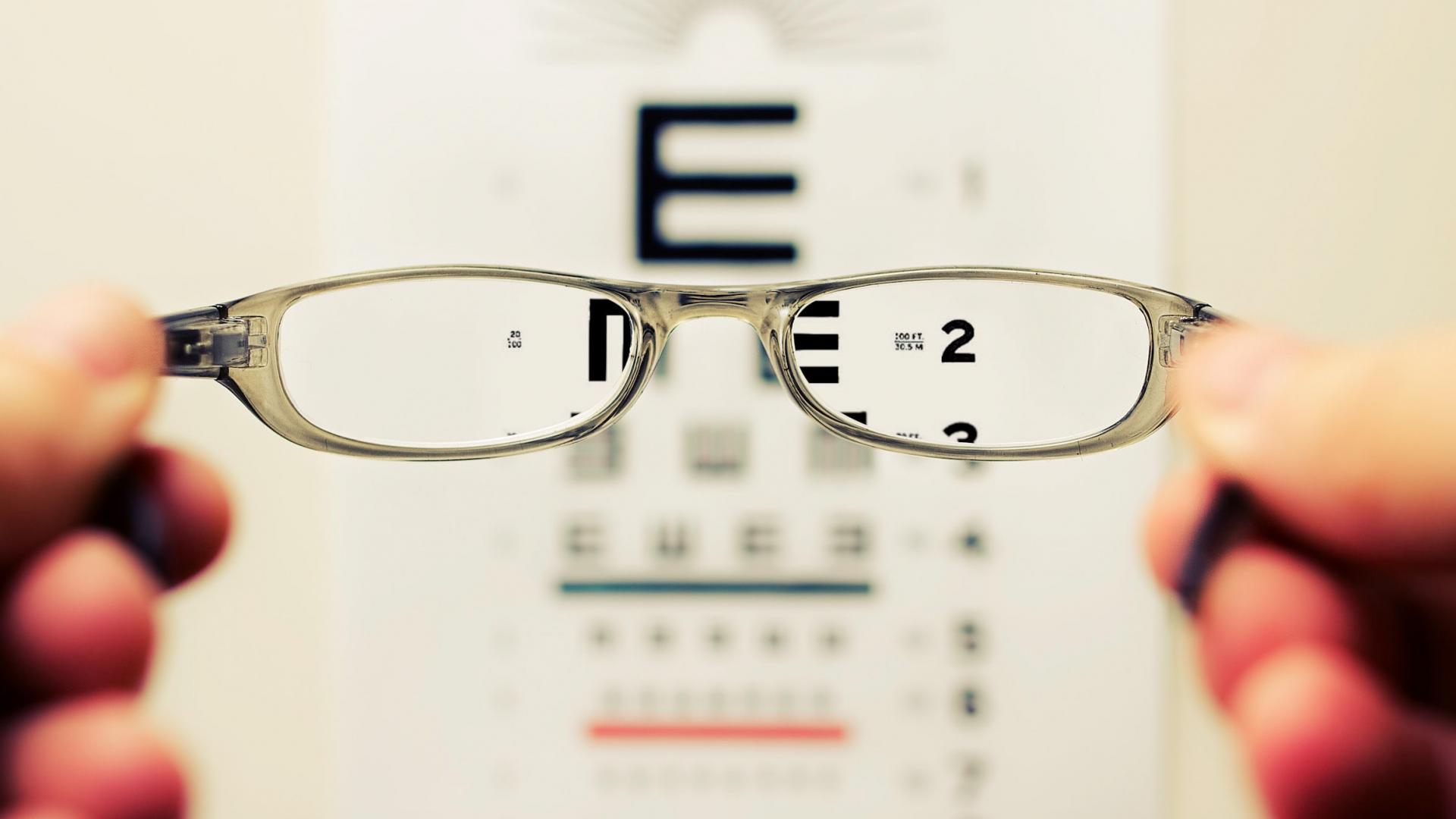 A man holding glasses in front of his face. The eye chart in the distance is in focus through the eyeglass lenses.