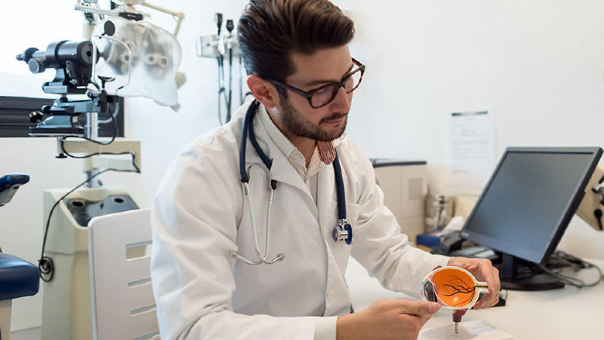 a man in a white doctor's coat examines a cross-section model of an eye