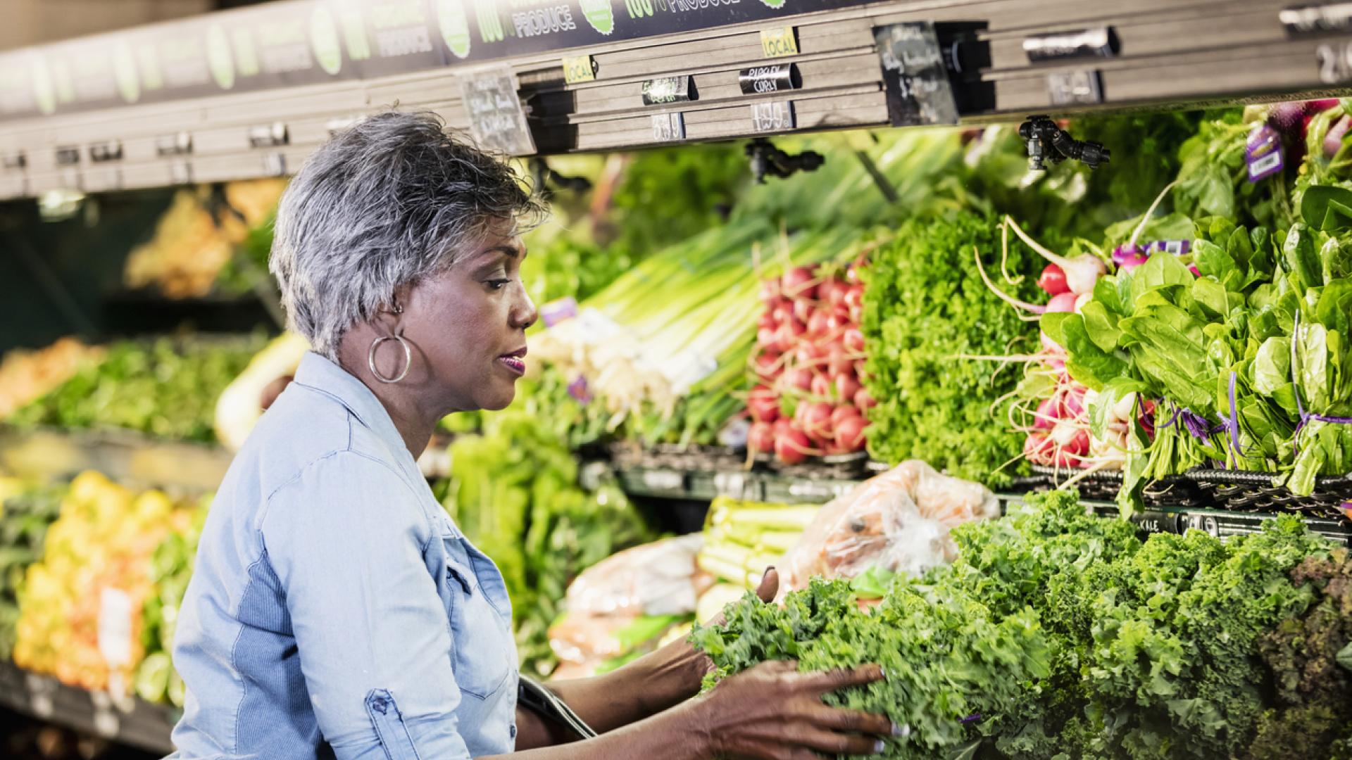 An older woman with short gray hair is shopping for fresh vegetables in a grocery store. She is wearing a light blue denim shirt and is selecting leafy greens from a well-stocked produce section filled with a variety of colorful vegetables, including radishes, spinach, and lettuce.