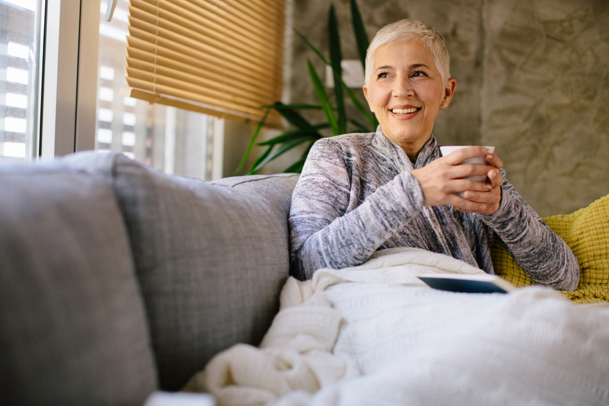 A person with short blonde hair smiling while holding a cup, sitting comfortably on a couch with cushions, near a window with blinds.