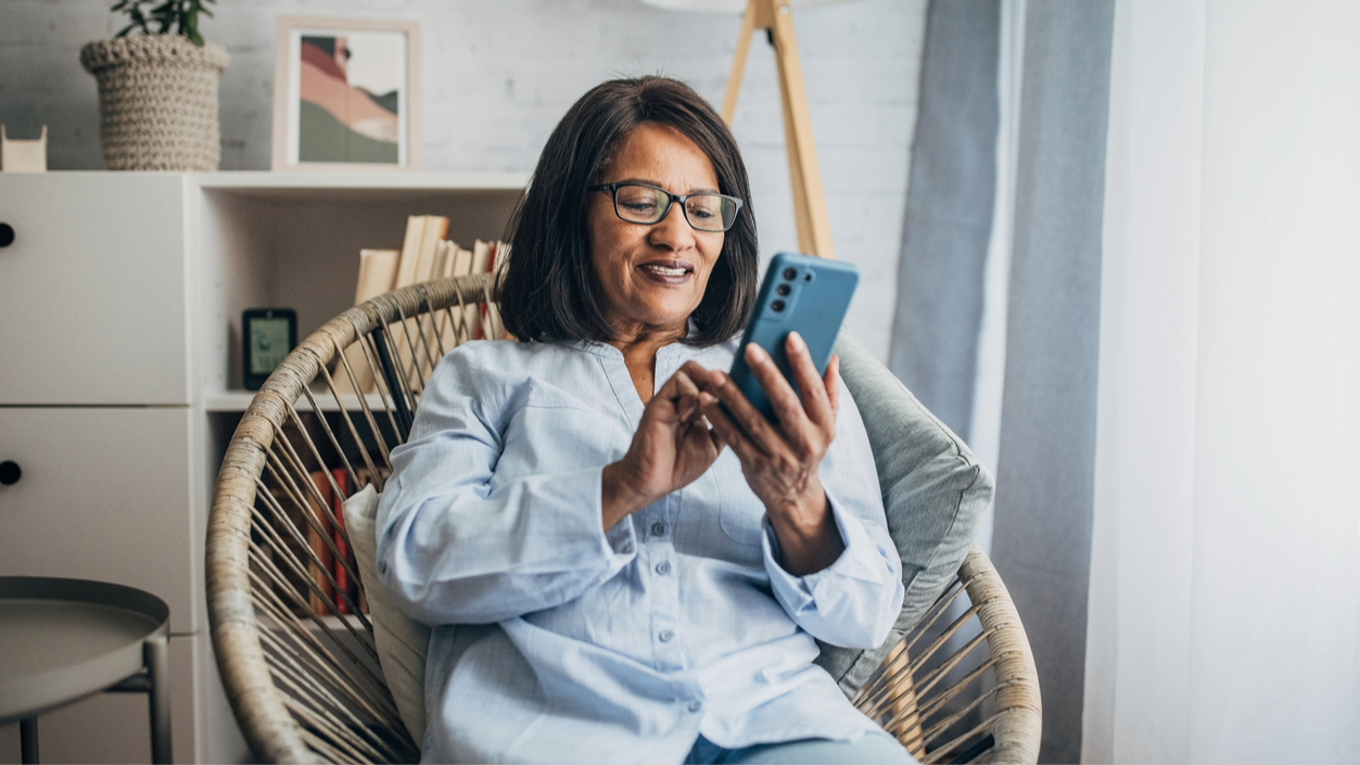 Person sitting in a wicker chair using a smartphone, smiling and appearing engaged, in a cozy room with a bookshelf and artwork in the background.