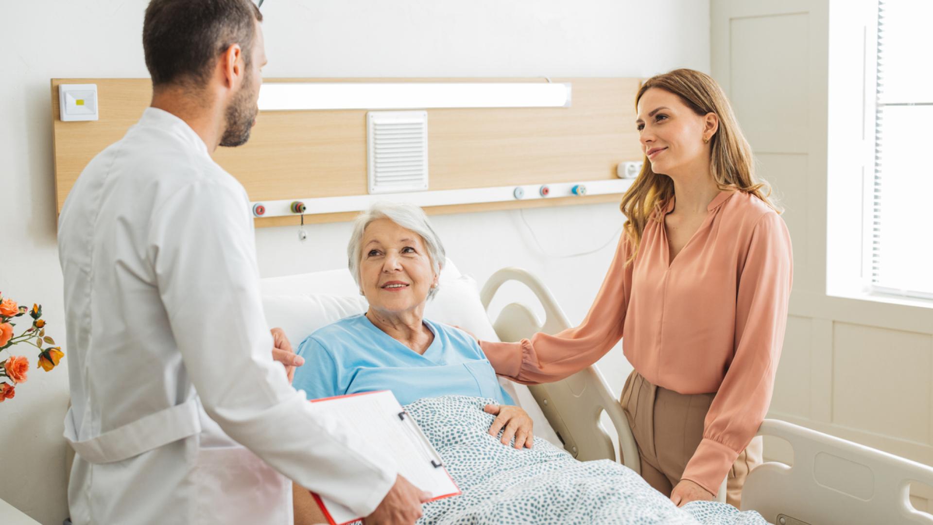 a doctor and a younger woman flank an older woman sitting, reclined, in a hospital bed. The older woman appears relaxed and is smiling.