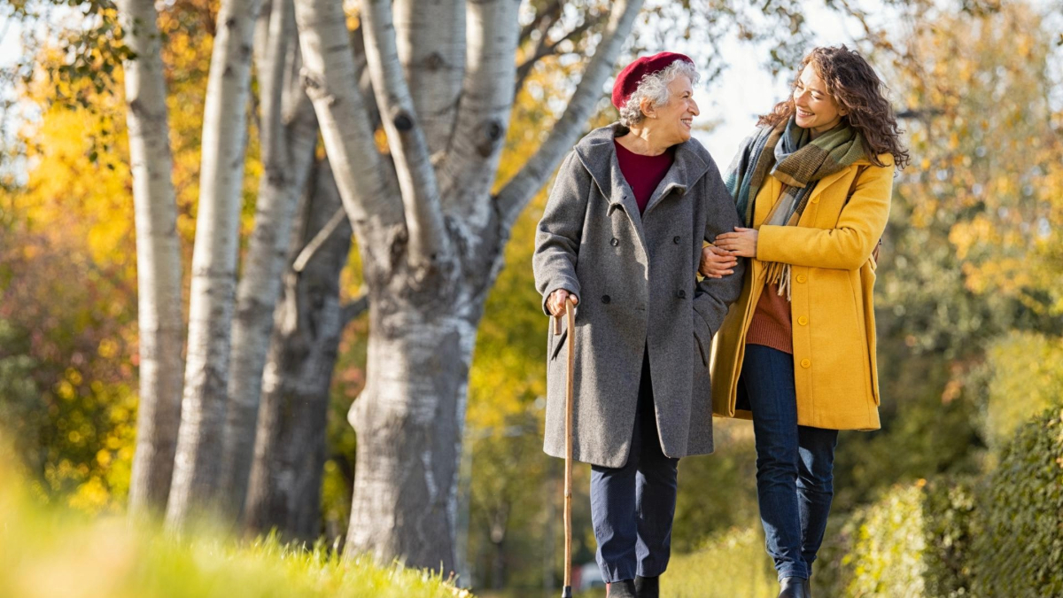 Two people walking arm in arm in a sunny park filled with autumn leaves, one older with a cane and one younger, both smiling and conversing.