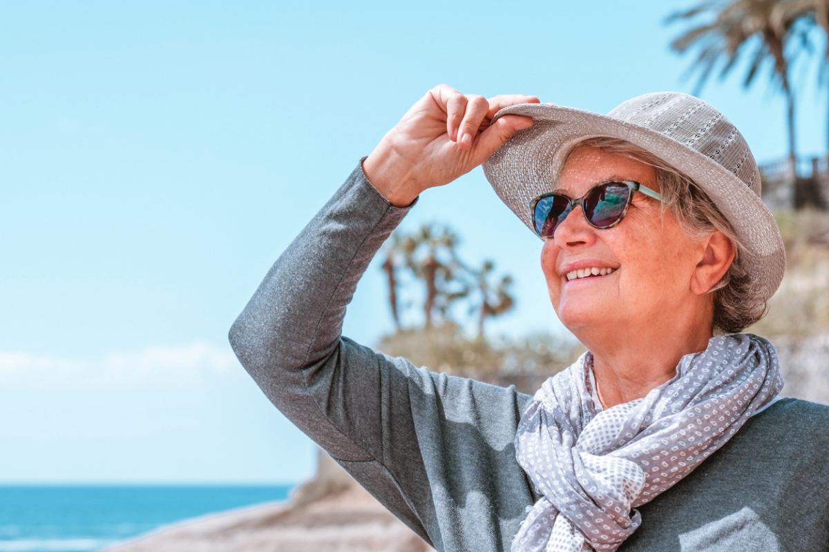 Person in a sun hat and sunglasses smiling on a sunny beach with palm trees in the background.