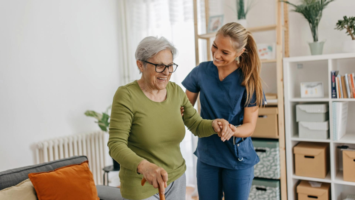 A caregiver assisting an elderly person with walking in a bright living room.