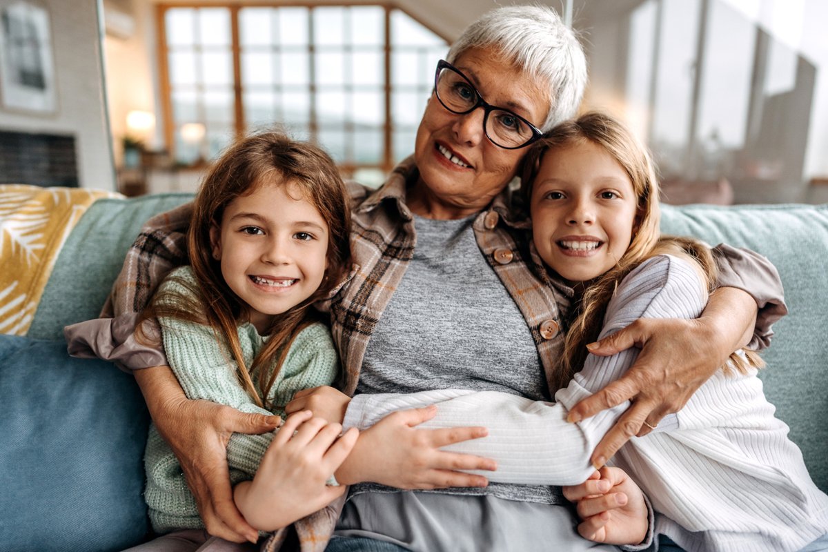 A woman sits on a couch hugging her granddaughters.