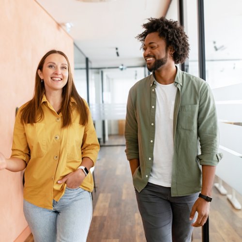 Two coworkers walk down the hall, smiling and laughing.