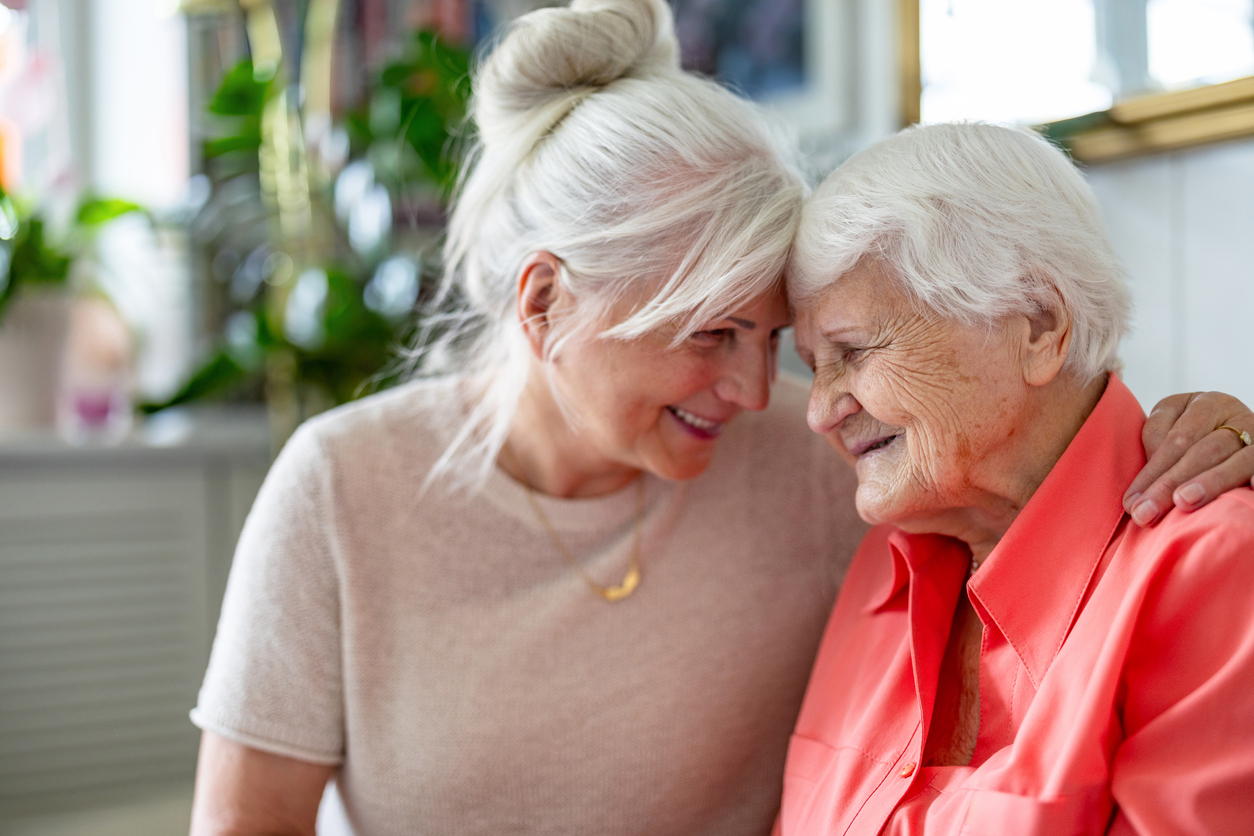 A heartwarming moment between an elderly woman and her adult daughter. Both have white hair, and they are smiling warmly at each other, with their foreheads touching in a loving gesture. The scene conveys tenderness and closeness, set in a bright, cozy indoor environment.