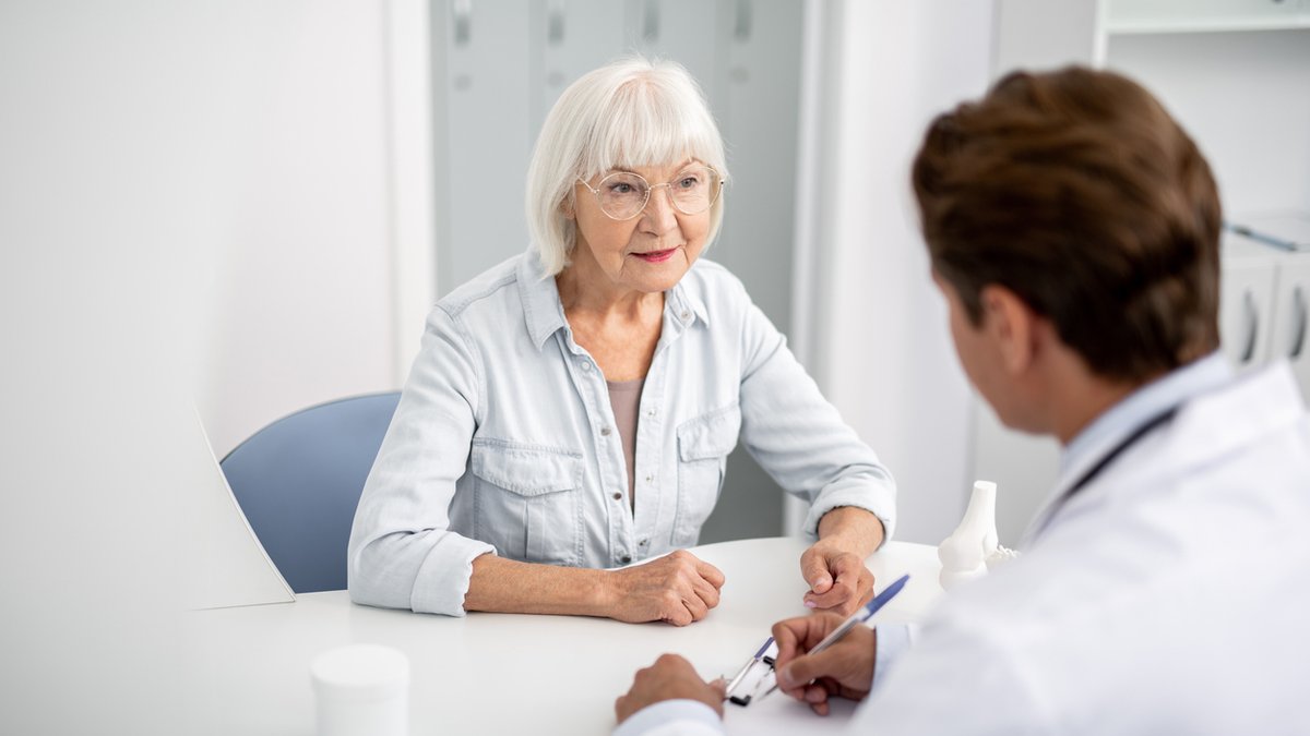 An elderly woman with white hair and glasses sits at a desk, attentively listening to a doctor during a consultation. The doctor, partially visible from the back, takes notes on a clipboard. The setting is a bright, clean medical office.