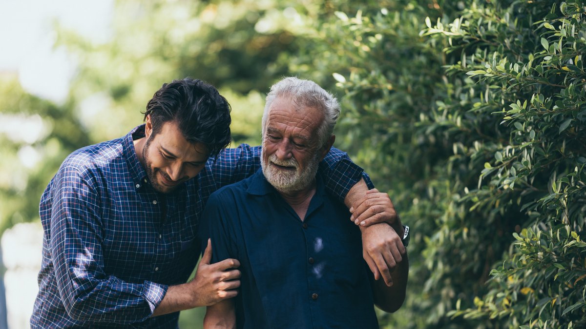 An older man with a white beard and short gray hair walks outdoors, supported by a younger man who has his arm around him. They are smiling and appear to be enjoying each other's company in a lush, green park setting.