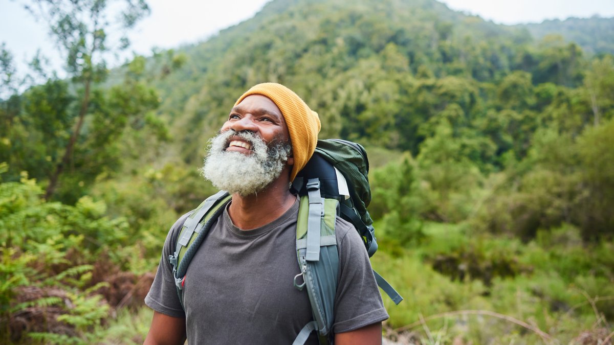 A bearded man with a gray beard and a yellow beanie smiles while looking up, standing outdoors with a backpack against a backdrop of lush, green mountainous landscape. He appears to be enjoying a nature hike in a serene forest setting.