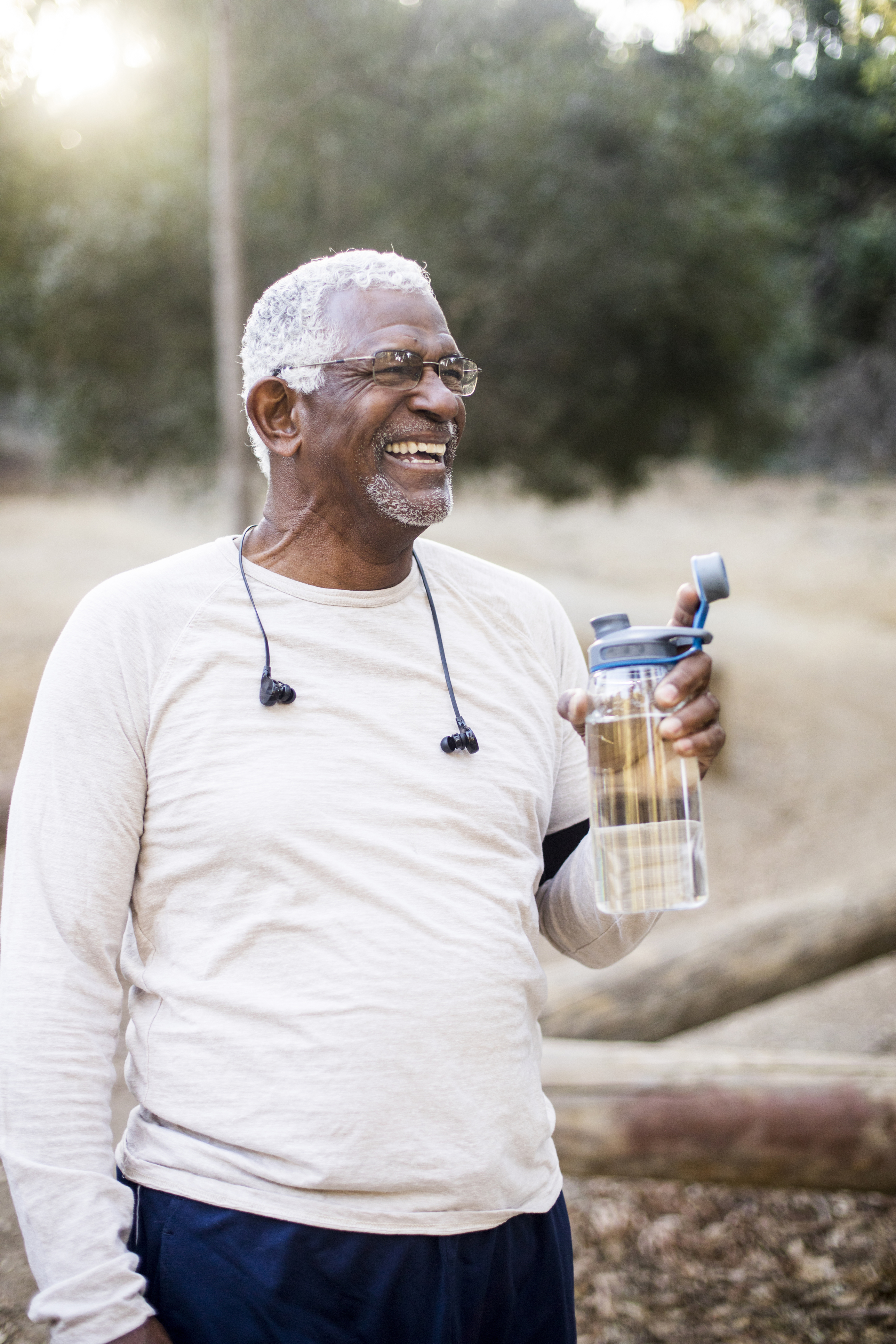 An older man with white hair and glasses smiles joyfully while holding a water bottle. He is outdoors, wearing a light beige long-sleeve shirt and earphones, with a natural background of trees and sunlight.