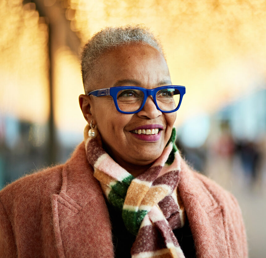 Head and shoulders view of mature individual with short gray hair wearing eyeglasses, coat and scarf, standing beneath glittering lights, looking away, smiling.